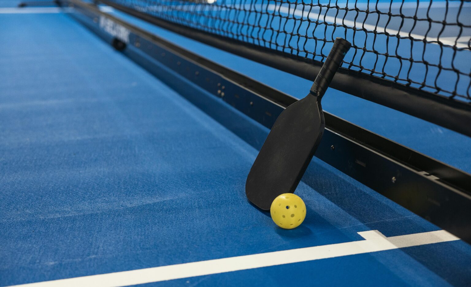 Pickleball paddle resting against net with yellow ball on blue court