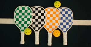 Four checkerboard-patterned pickleball paddles with wiffle balls on a sports court.