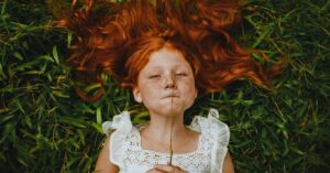 A young redhead girl lies in the grass holding a blade of grass, showcasing her freckles and peaceful expression.