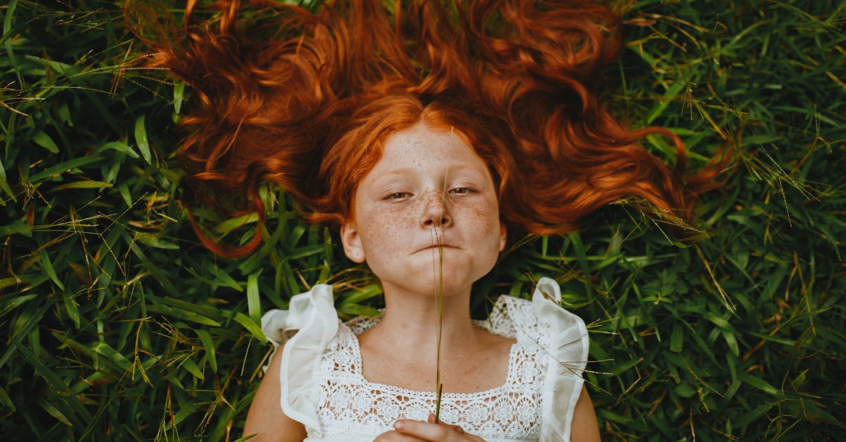 A young redhead girl lies in the grass holding a blade of grass, showcasing her freckles and peaceful expression.