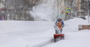 Person using a snow blower to clear a city sidewalk during a heavy winter snowfall.